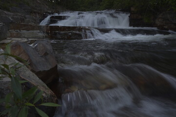 Flowing creek water over rocks with waterfall in background. Green plant in lower left corner