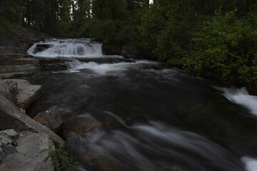 Flowing creek water over rocks with waterfall in background along with green foliage.