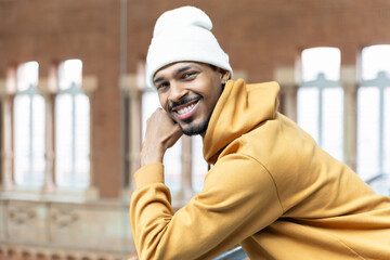 Portrait of smiling hispanic man leaning on railing, wearing beanie and hoodie