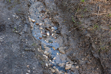 A muddy dirt road heavily eroded by a small stream. Water flows through a shallow channel filled with stones and broken bricks. Top-down view showing details of erosion and debris.