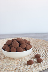 Chocolate-coated almonds overflowing white ceramic bowl, surrounding scattered nuts on textured woven surface
