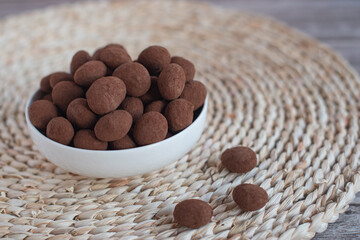 Chocolate-covered almonds overflowing a white bowl on a woven placemat, creating a tempting snack or dessert