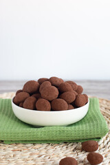 Chocolate covered almonds overflowing a white bowl on a green napkin and wicker place mat, making a sweet treat