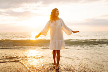 A joyful woman in a white dress has fun on the sunset beach. Young woman enjoying the sea and...
