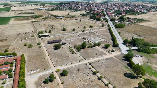 Aerial view of an archaeological site of Dion, Greece with ruins, surrounded by dry fields and a small village in the background. The landscape features sparse trees and a road nearby.