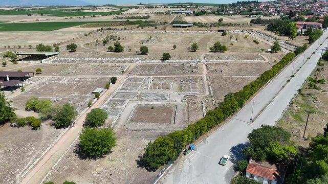 Aerial view of an archaeological site of Dion, Greece with ruins, surrounded by dry fields and a small village in the background. The landscape features sparse trees and a road nearby.
