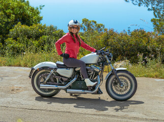 Obraz premium Beautiful woman smiling looking at camera wearing an open face helmet sitting posing on her vintage Harley-Davidson Sportster motorcycle outdoors on a sunny coastal road.