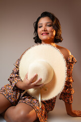Attractive brown skin woman sitting on the ground, smiling and holding a summer straw hat. Perfect for beauty, fashion, lifestyle, vacations, femininity. Vertical studio portrait with copy space.