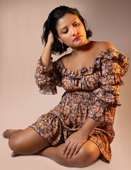 Charming brown skin woman wearing  floral pattern dress, sitting on the ground and placing her hand in hair. Perfect for beauty, lifestyle, femininity. Vertical studio portrait with copy space.