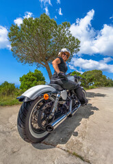 Obraz premium Confident woman smiling at the camera dressed in biker clothing sitting on her custom chopper motorcycle under a blue sky on a sunny coastal road.