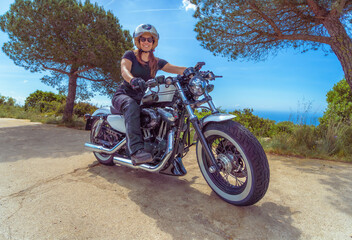 Smiling young woman enjoying riding her Harley-Davidson motorcycle on a sunny coastal road with the sea in the background.