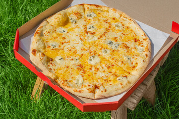 Cheese pizza in a red box on a stool on the grass. Food delivery, pizza.