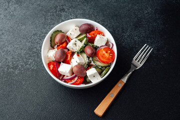 Greek salad with feta cheese in white bowl with fork on dark background