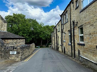 A road lined with stone cottages winds past Edroyd Place, opening into lush greenery under a partly cloudy sky—serene countryside charm in Farsley, Leeds, Yorkshire, UK.
