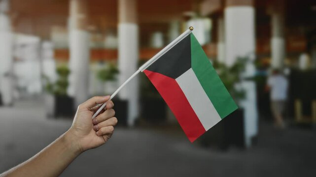 Male hand holding kuwait flag in vibrant outdoor city setting displaying national pride and multiculturalism among a diverse urban backdrop of a bustling town.