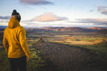 Hiker in a yellow jacket stands on top of Midfell in Hrunamannahreppur, Iceland, taking in the golden valley view at sunset. A quiet moment of reflection and wonder. © Fauren