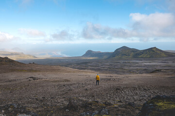 Lone traveler in a yellow jacket walks across Iceland’s volcanic highlands toward the distant coastline, surrounded by raw beauty and the vastness of Snafellsnes Peninsula