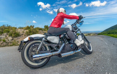 Obraz premium Side view of a female motorcyclist wearing a red jacket, tights, and white boots, riding her customized Harley-Davidson chopper motorcycle taking a hairpin turn along a scenic mountain road.