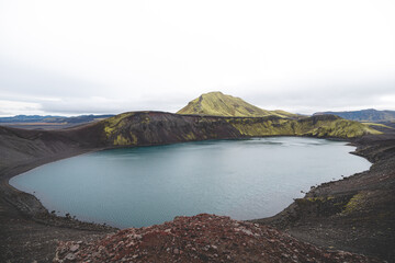 Serene volcanic lake with a tiny mossy island sits in the Icelandic highlands, surrounded by rugged hills and untouched nature under a soft overcast sky. Highlands in central Iceland