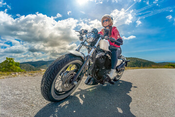 Obraz premium Frontal view from below of a smiling woman on a custom chopper motorcycle, driving around a curve under a sunny sky with mountains in the background.