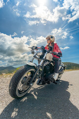 Fototapeta premium A young woman enjoying riding a classic chopper motorcycle looking at the mountains on a mountain road under a sunny sky with clouds illuminated by the sun.