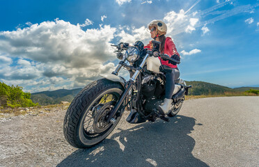 Obraz premium Smiling woman wearing a red jacket enjoying a ride on her customized chopper motorcycle on a mountain road, looking at the panoramic horizon under a sunny sky with mountains in the background.