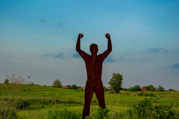 A tall metal sculpture depicts a figure with raised hands against a colorful sky. Green fields...