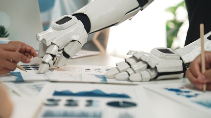 A close-up view of a robotic arm working alongside human hands in a collaborative business meeting, highlighting the integration of technology and strategy in modern workplaces.Noogenesis