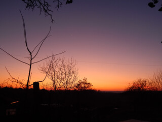 A stunning sunset paints the sky in a dramatic gradient from purple to fiery orange, silhouetting bare trees and a distant horizon, with faint stars beginning to appear.