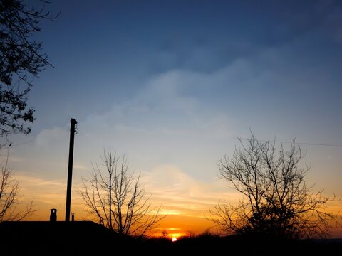  A striking sunset with a vibrant orange glow on the horizon, silhouetting bare trees and a tall, thin structure against a gradient blue sky.