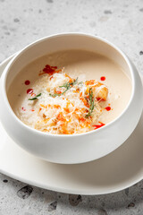 A bowl of creamy soup garnished with shredded cheese, herbs, and a drizzle of red sauce, presented on a white plate atop a textured gray surface. The dish is appetizing and elegantly plated