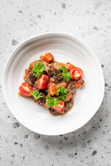 plate of homemade beef stew with tender chunks of meat, carrots, and cherry tomatoes, garnished with fresh parsley. Rich sauce, hearty comfort food, top view on a gray countertop.