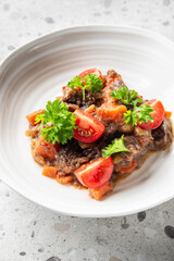 plate of homemade beef stew with tender chunks of meat, carrots, and cherry tomatoes, garnished with fresh parsley. Rich sauce, hearty comfort food, top view on a gray countertop.