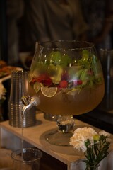 A large glass punch bowl with a tap filled with a refreshing drink with pieces of fruit (such as lime, strawberry or raspberry) and mint leaves.