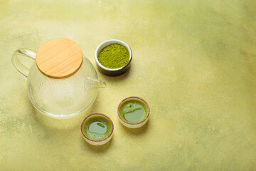 Steaming glass teapot with wooden lid beside two small cups of matcha tea and a bowl of matcha powder on a textured green background, evoking calm and wellness, copy space