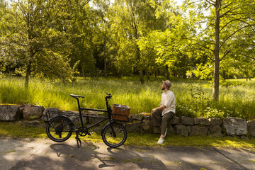 Caucasian man sitting in park with cargo bicycle on a sunny day wearing a hat.