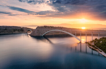 Aerial view of bridge and blue sea at colorful sunset. Modern Krk bridge island during golden hour, mountains, rocks, reflection in water and orange sky. Top drone view of bridges. Architecture