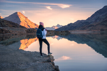 Young woman hiker with backpack standing on the shore of a mountain lake enjoying the sunrise over the swiss alps in autumn. Concept of adventure, travel, healthy lifestyle. Girl on the lake in fall