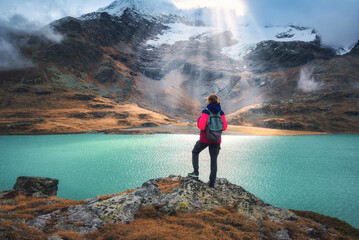 Young woman hiker with backpack standing on a rock with view on turquoise alpine lake, sunbeams, snowy mountain and glacier in the background in fall. Girl on mountain trail in Swiss Alps in autumn