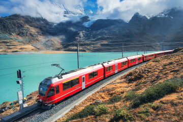 Red train is moving along railway in swiss alps mountain range near lake on a sunny day with cloudy sky in autumn. Landscape. Bernina Express train, orange grass, snowy rocks at sunset in fall. Travel