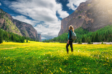 Girl with backpack on the meadow with yellow flowers and green grass in beautiful alpine mountain valley at sunrise in summer.Young woman in alps, sky with clouds. Hiking. Spring in Dolomites, Italy