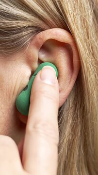 Extreme close-up macro video of a human finger gently inserting a bright green foam earplug into an adult ear canal, showing skin texture and blurred blonde hair. Concept for health and wellness