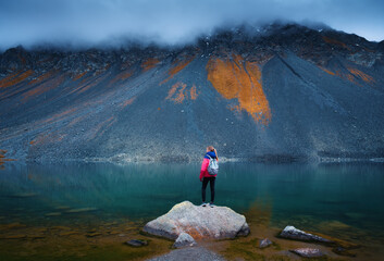 Young woman hiker with backpack on a rock in autumn. Girl and breathtaking view of a mountain alpine lake reflecting the surrounding peaks, orange grass and cloudy dramatic sky. Switzerland in fall