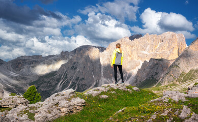 Girl on the hill with yellow flowers and green grass in beautiful alpine mountain valley at sunset in summer. Landscape with young woman in alps, sky with clouds, rocks. Travel in Dolomites, Italy