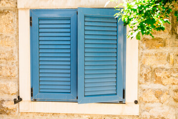 Closed blue window shutters on stone wall with bright summer light. Mediterranean facade, architectural detail, and urban tranquility.