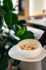 A creamy soup in a white bowl, garnished with fresh herbs and spices, served on a plate. The background features a green plant and a blurred indoor setting, suggesting a cozy dining atmosphere