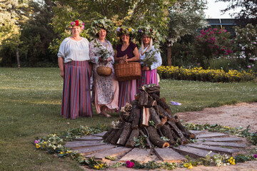 On the evening of the summer solstice, women stand with wreaths on their heads, baskets and flowers in their hands, and colorful clothes. Outdoors, by a campfire.