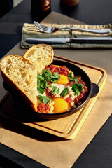 Shakshuka with coriander and mint served with bread in a black pan on a wooden board