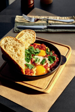 Shakshuka with coriander and mint served with bread in a black pan on a wooden board