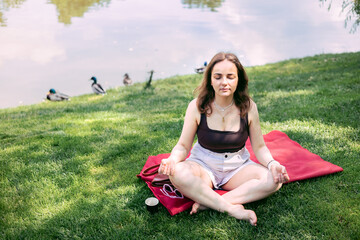 Healthy and beautiful young woman practicing yoga in the park, sitting on grass, meditating with closed eyes near lake.